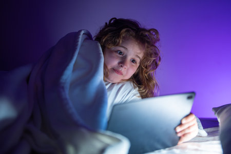Kids Bed time story in tablet. Kid lying on bed and surfing Internet on tablet in dark room. Boy reading Tablet in bed at night. E-book, story and kids app game. Kid using tablet before going to bed.の写真素材