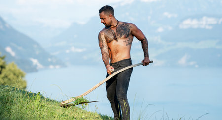 Strong farmer using scythe. Sexy Farmer with a scythe on green grass field. Muscular Farmer cut grass. Rural farmer on summer meadow mowing grass. Meadow lawn cutting. Summer farm. Hay making.の写真素材