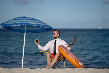 Successful businessman on summer vacations. Funny business man drink summer cocktail and using laptop in sea beach. Summer business concept. Hot summer. Crazy work. Remote online working.の写真素材