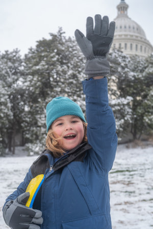 Portrait of kid in winter Washington DC. Child in knitted hat playing in a snowy winter park near Capitol. Child playing with sleigh ride. Winter kid play in snowy city park. Winter vacation with child.の写真素材