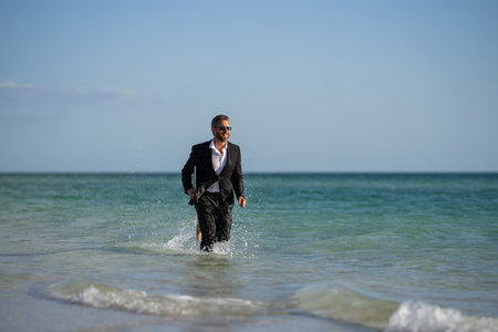 Freelance work, online business. Summer business. Businessman in suit hold laptop and run on sea. Funny business man in a business suit with laptop standing in water. Remote work. Crazy freelancer.の写真素材