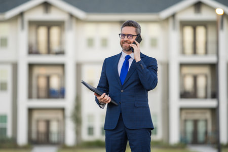 Real estate agent business man in suit hold holder clipboards and talking on phone. Businessman. Business Man broker or real estate agent. Business man in suit outdoor. Real estate business man.の写真素材