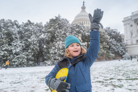 Portrait of kid in winter Washington DC. Child in knitted hat playing in a snowy winter park near Capitol. Child playing with sleigh ride. Winter kid play in snowy city park. Winter vacation with child.の写真素材