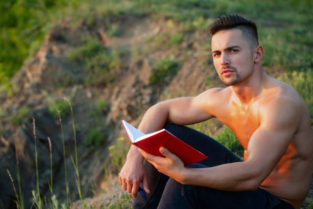 Sexy romantic man with naked body reading a book outdoor. A handsome intellectual. Nerd or smart male model with sexy body reading book. Man holding a book on the grass in the park.の写真素材