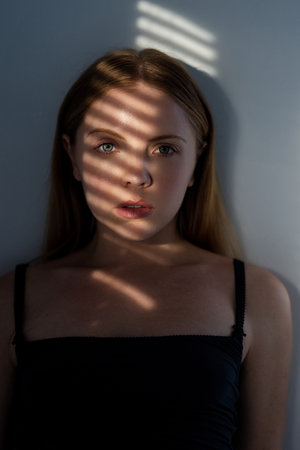 Woman looking through window blinds. Portrait of a woman in shadows on face from window blinds. Girl pensive look at window in living room. Portrait female model with shadow from the blinds.の写真素材