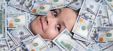 Kid with fun face in money. Kid peeking out of dollar bills with astonished shocked eyes. Money banknotes, cash dollars bills. Money background.の写真素材