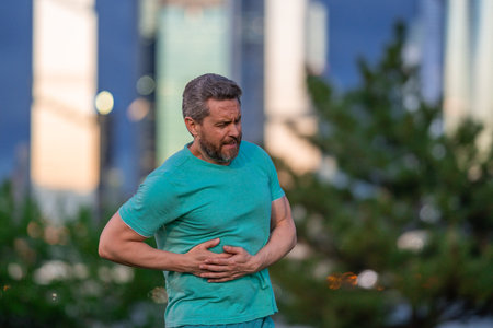 A man having a heart attack outdoors. A man clutching his chest in pain. A man from a heart attack in a park. A man experiencing chest pain while walking.の写真素材