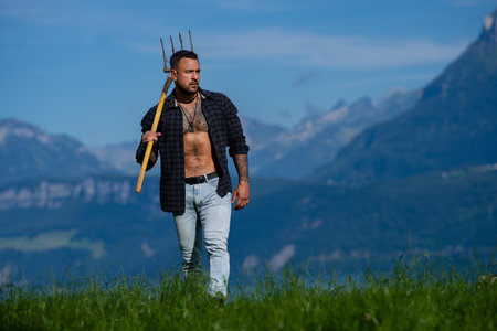 Rural strong man working on a farm. Farmer working in a field under the sun. Farmer with pitchfork in a grassy field. Rural laborer in a green field. Hardworking man in rural countryside.の写真素材