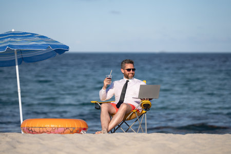 Successful businessman on summer vacations. Funny business man drink summer cocktail and using laptop in sea beach. Summer business concept. Hot summer. Remote online working.の写真素材