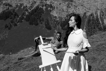 Romantic Artists couple drawing outdoor. Sensual couple artists draw pictures in alps mountain. Woman painting with brushes oil or acrylic paint. Man teaches to draw a girl.の写真素材
