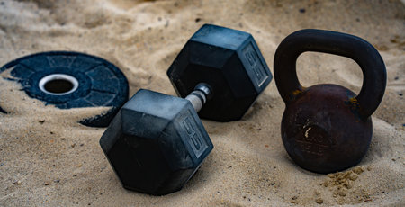 Outdoor gym background. Metal kettlebell on sand. Natural workout. Gym weights background. Summer training background. Barbell and dumbbell background.の写真素材