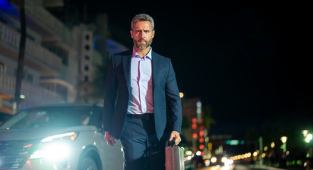 Business man with briefcase in suit walking at night city. Rich man in a suit outdoor. Mature business man walk in night city lights. Business style. Wealthy businessman.の写真素材