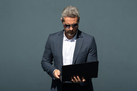 Businessman working on a laptop. Confident man using a laptop in a city. Executive in a suit typing on a laptop. Successful entrepreneur working remotely. Business man checking emails on a laptop.の写真素材