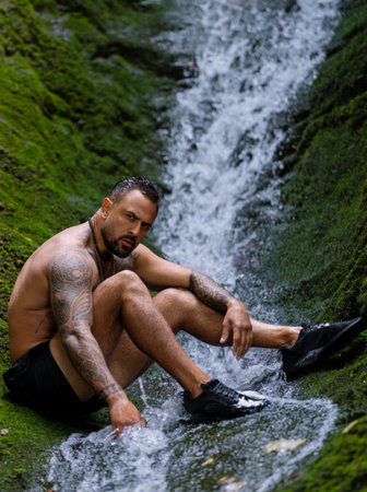 Shirtless man splashing under waterfall. Fit and strong body. Hispanic man enjoying summer water....