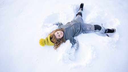 Child making a snow angel. Happy kid lying in the snow. Kid angel in snow. Winter kids with snowy angel wings. Smiling child on a snowy day. Carefree kid in the snow lake angel.の写真素材