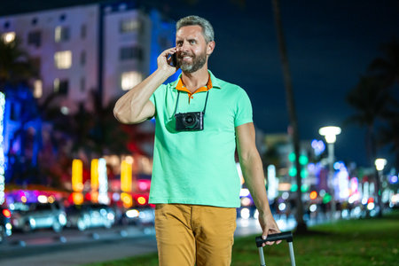Man talk on phone in night street. Tourist calling on phone in night city. Hispanic guy using phone. Mature man talking on phone during travel. Handsome mature man in t-shirt with cellphone.の写真素材