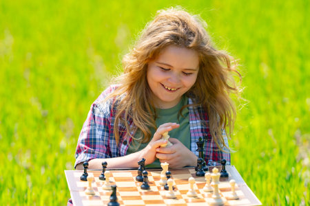 Kid plays chess. A clever cute child thinks and playing chess. Kid focuses on chessboard. The child make checkmate. A smart kid plans chess. Child sits on grass enjoys chess. The board logic game.の写真素材