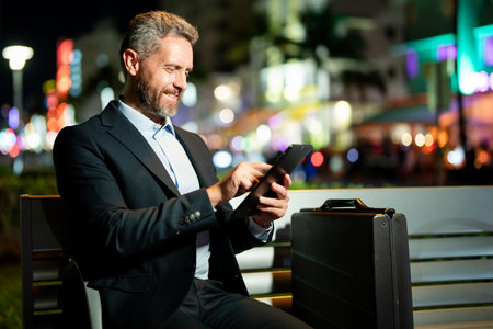 Online night business. Businessman working on tablet outdoors. Businessman with tablet outdoor. Businessman with a tablet on night street. Businessman using tablet, sit on bench. Night business.の写真素材