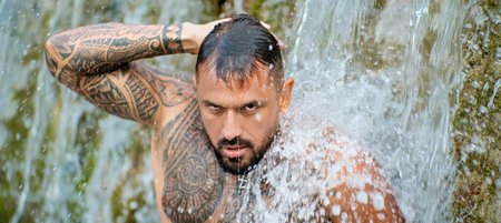 Shirtless man splashing under waterfall. Fit and strong body. Handsome model enjoying summer water. Athletic male body. Man beauty on nature. Muscular body with water drops.の写真素材