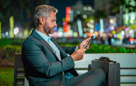 Online night business. Businessman working on tablet outdoors. Businessman with tablet outdoor. Businessman with a tablet on night street. Businessman using tablet, sit on bench. Night business.の写真素材