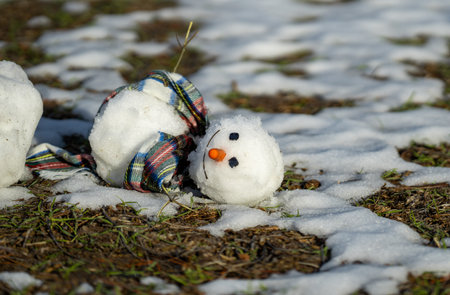 Snowman is melting outdoors in sunlight on snowy background mixed with green grass. Approaching spring, warm winter, climate change concept. A melted snowman as symbol of the end of the winter.の写真素材