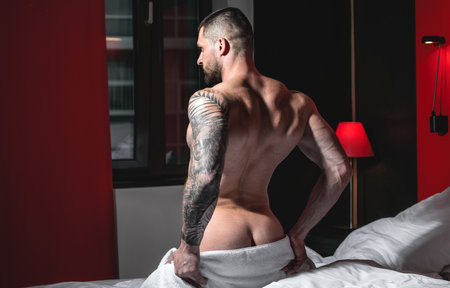 Handsome nude man lying in a bed. Top view of handsome young man in bed in bedroom at night. Muscular handsome man laying on stomach in bed. Guy lying on belly and covered sexy body by blanket.の写真素材