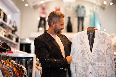 Suit male fashion shop. Man shopping fashion jacket. Stylish man in suits boutique. Mature man choosing jacket. Designer jacket. Glamour jacket with rhinestones in luxury store. Fashionable clothes.の写真素材