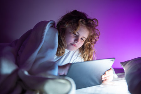 Kid lying on bed and surfing Internet on tablet in dark room. Boy reading Tablet in bed at night. Kid in pajamas using tablet before going to bed. Kid using tablet gadgets.の写真素材