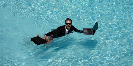 Business man in suit working on laptop in swimming pool. Travel tourism and business concept. Male office employee using laptop in pool on summer day.の写真素材