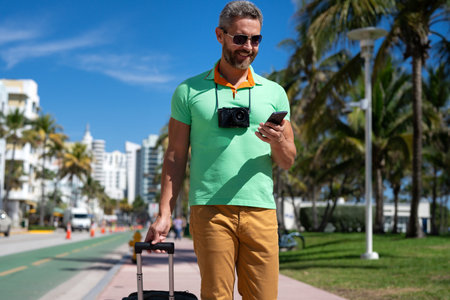A traveler with camera and luggage on streets of Miami. A man traveler enjoys his summer vacation in a tropical city. A traveler tourist with suitcase on summer city. Man traveler walk on city.の写真素材