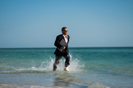 Businessman suit barefoot running on beach. Handsome business man on summer vacation. Businessman running on sea holding laptop. Businessman excited jumping in sea water. Crazy businessman.の写真素材
