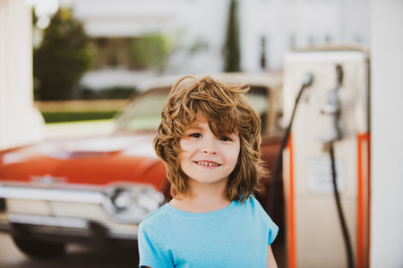 Kid holding fuel nozzle during road trip. Portrait of little traveler with gasoline pump. Childhood vacation moment at petrol station. Boy near vintage car and fuel tank. Travel memories with child and auto.の写真素材