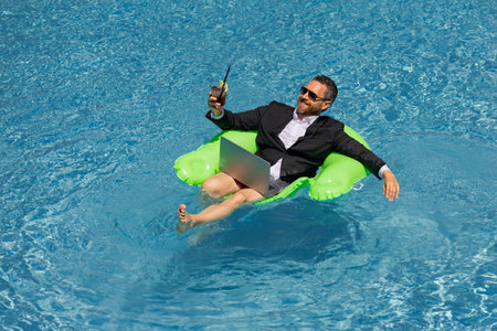 Crazy summer. Business drink summer cocktail and using laptop in suit in water pool. Fun business lifestyle. Funny middle aged business man dreams about summer. Summer travel. Business success.の写真素材