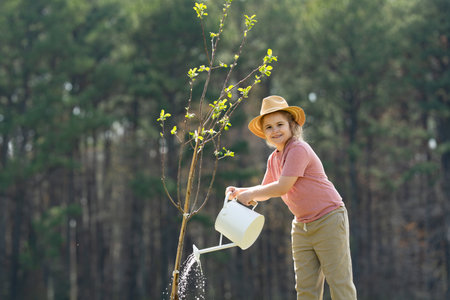 Child in hat cultivating tree with shovel. Child cultivating tree on backyard. Child cultivating tree in spring field. Little gardener with watering can and shovel. Child cultivating tree in garden.の写真素材