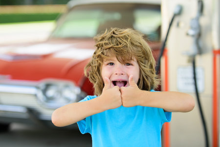 Happy child refueling retro car at gas station. Kid holding fuel nozzle during road trip. Portrait of little traveler with gasoline pump. Kid on road trip. Classic car and kid at old-style fuel station.の写真素材