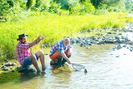 Young and old fisherman standing on the shore of lake with fishing rod. Father and son enjoy life. Men family, granddad and drandson fishing. Mature man fisher celebrate retirement.の写真素材