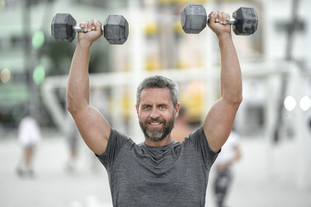 Sporty man lifting dumbbells outdoors. Athletic male exercising with dumbbells in public outdoor gym. Fit guy working out in the park. Sportsman lifting dumbbells. Mature man training with dumbbells.の写真素材