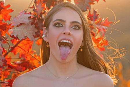 Young woman collects yellow fall leaves in autumn. Woman holds near autumn yellow leaf with tongue. Portrait of sensual woman over autumn fall.の写真素材