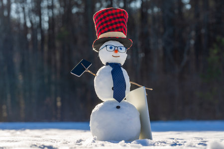 Snowman talking on smartphone in snowy landscape. Snowman making a phone call. Funny snowman using laptop outdoors. Snowman with mobile phone during winter celebration.の写真素材