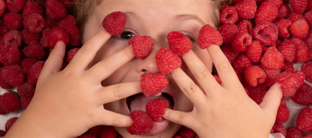 Funny kids face near raspberry background. Cute child eats raspberries from fingers. Child face with raspberry frame, close up raspberry background.の写真素材