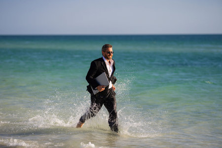 Summer business. Freelancer run on summer sea beach. Businessman in wet suit jump in sea water. Crazy business summer vacation. Funny businessman with laptop in suit on beach. Hot summer.の写真素材