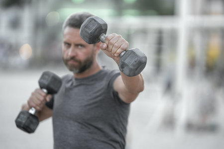 Dumbbells in arm. Closeup of a mature man lifting dumbbells outdoors. Man arm muscles engaged in a...