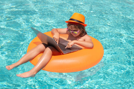Happy little boy learning online computer laptop. Kid relax on floating ring using computer in swimming pool. Summer business and technology.の写真素材