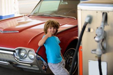 Happy child refueling retro car at gas station. Kid holding fuel nozzle during road trip. Portrait of little traveler with gasoline pump. Kid on road trip. Classic car and kid at old-style fuel station.の写真素材