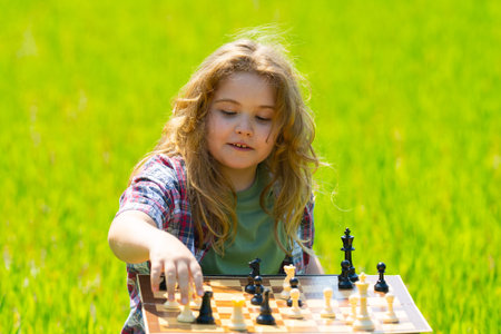 Kid plays chess. A child thinks and playing chess. Kid focuses on chessboard. The child make checkmate. A smart kid plans chess. Child sits on grass enjoys chess. Board logic game for child.の写真素材