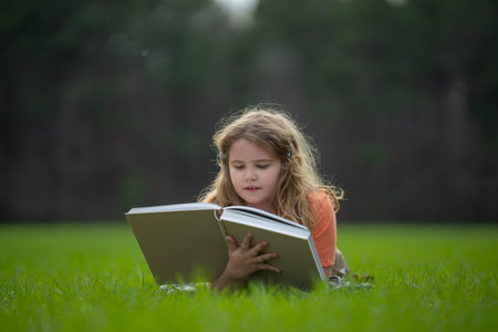Child reading a book in the grass. Child enjoying a book story outdoors. Child studying with book in the meadow on a sunny day. Learning in nature. Schoolboy reading book. Child outdoor learning.の写真素材