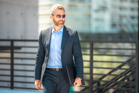 Successful business man walk with on laptop outdoors. Confident business man hold laptop outside. Mature freelancer businessman in suit. Corporate business. Rich business man in suit.の写真素材