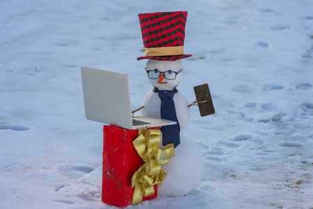 Businessman snowman talking on smartphone in winter. Snowman making a business phone call. Funny snowman hacker using laptop outdoors. Snowman with mobile phone during winter holiday.の写真素材