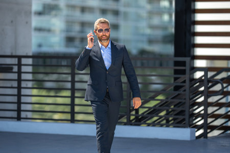 Businessman walking and talking on phone. Confident business man outdoors with phone. Business phone conversation. Busy corporate leader talking on phone. Rich business man in suit. Business call.の写真素材