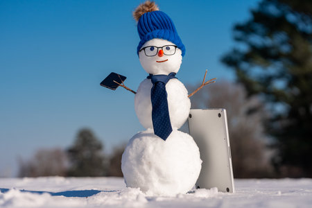 Businessman snowman talking on smartphone in snow. Snowman making a business phone call. Funny snowman hacker using laptop outdoors. Snowman with mobile phone during winter holiday.の写真素材